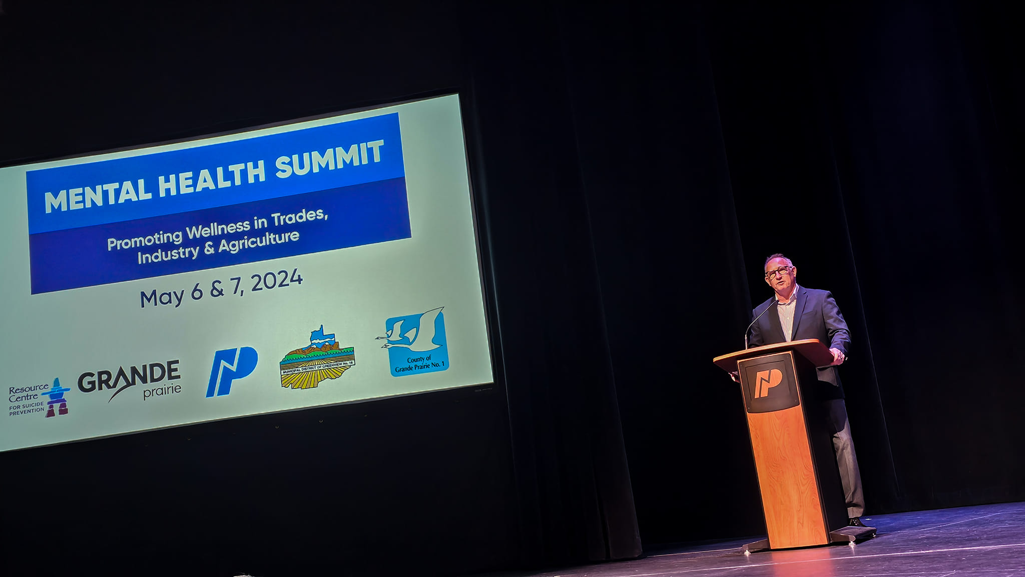 A speaker at a podium on stage beside a Mental Health Summit slide reading Promoting Wellness in Trades, Industry & Agriculture, May 6 and 7, 2024