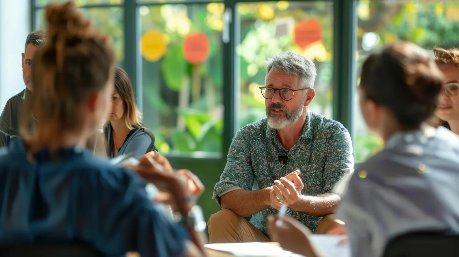 A man with grey hair and glasses speaking and gesturing during a group discussion around a table