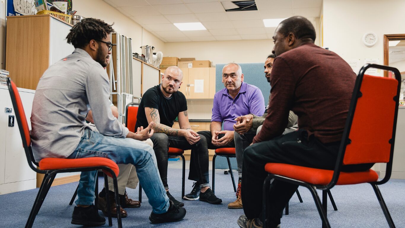 Six men seated in a circle of chairs in a meeting room, talking