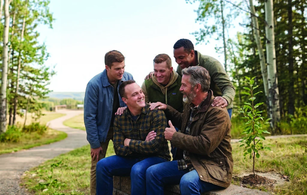 Five men gathered outdoors on a trail, one seated on a log bench with the others around him