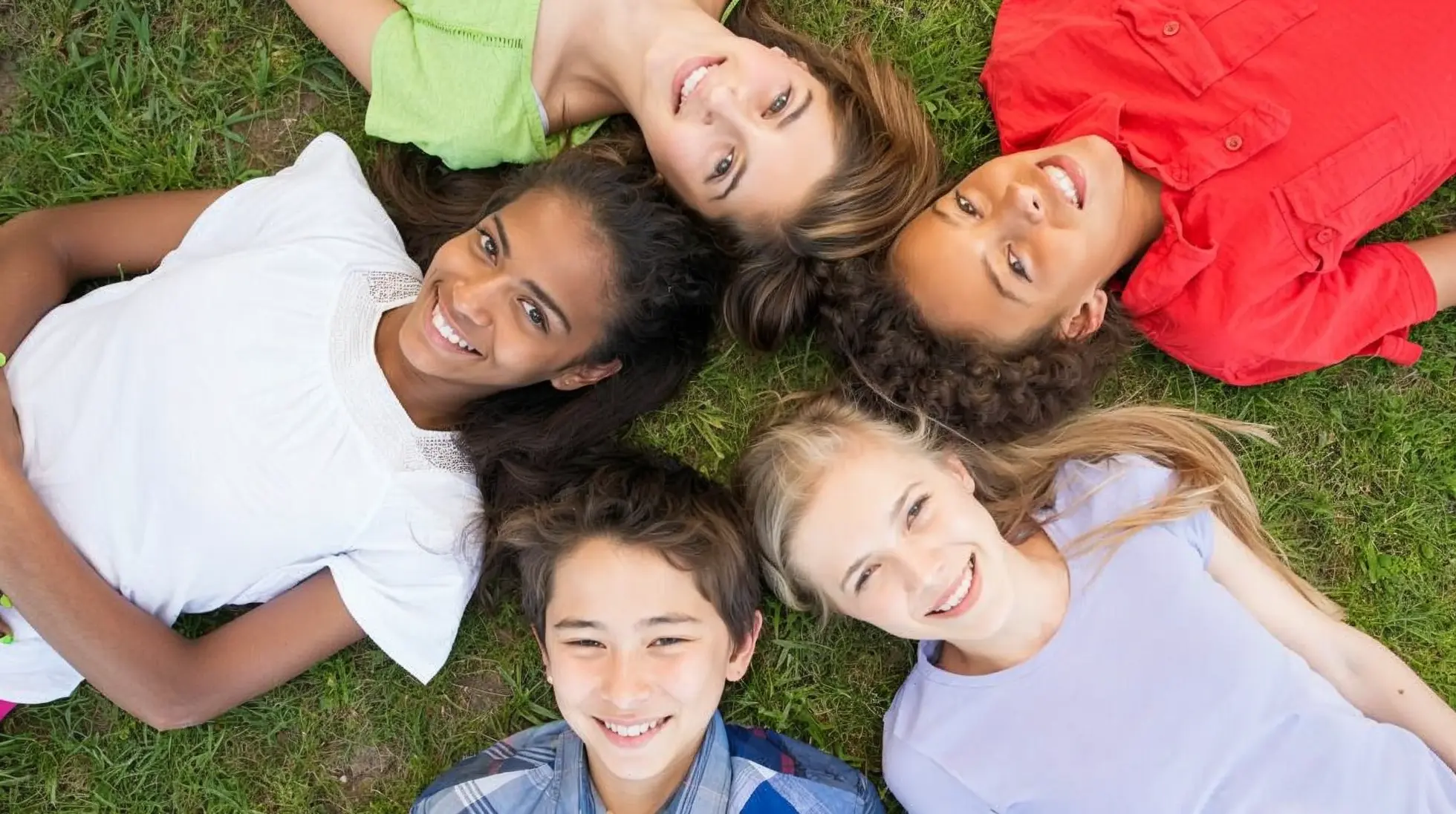 Five smiling young people lying on the grass in a circle with their heads together