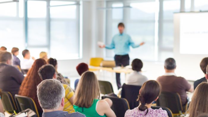A presenter speaking to a seated audience in a bright training room